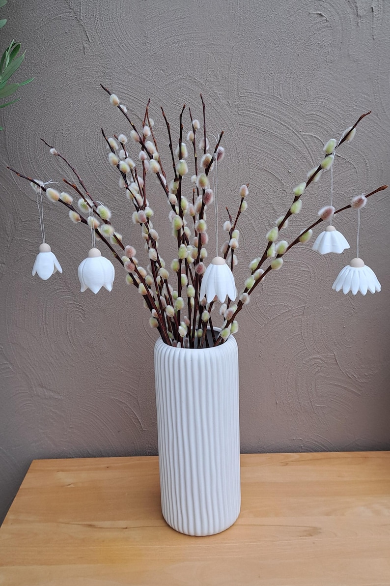 May include: A white, ribbed ceramic vase holds pussy willow branches with hanging white flower ornaments. The arrangement sits on a wooden surface against a textured brown wall. Decorative and seasonal.