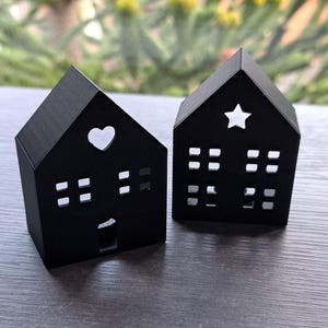 Puede incluir: Dos portavelas decorativos negros con forma de casa. Uno tiene un recorte en forma de corazón, el otro una estrella. Ambos tienen recortes de ventanas cuadradas. Las casas están sobre una superficie oscura con un fondo verde borroso.