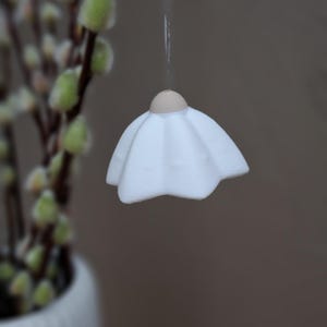 May include: A white, petal-shaped ornament hangs from a string. The ornament has a light brown top and a ruffled, flower-like design. The background is blurred, with a branch of pussy willow and the text "Typ 2" visible.