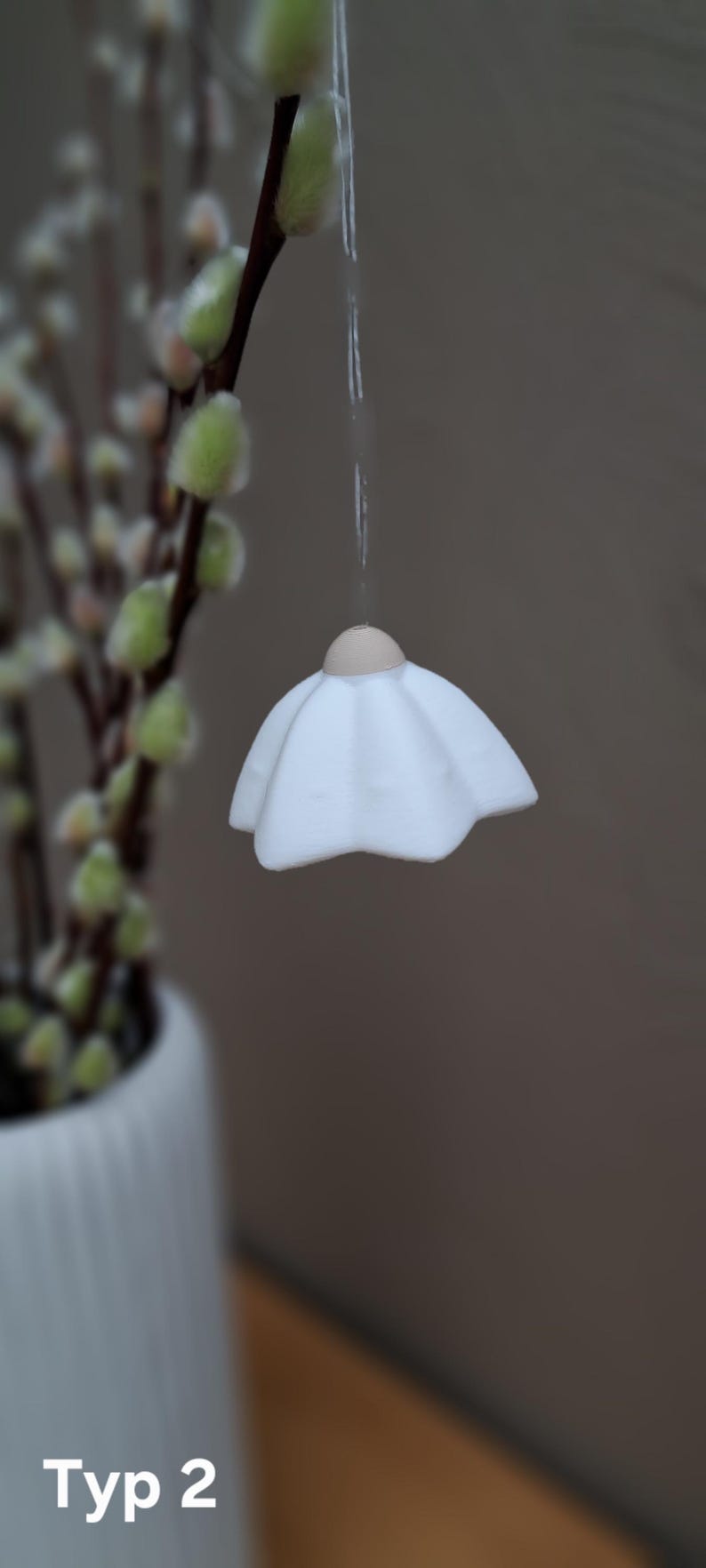 May include: A white, petal-shaped ornament hangs from a string. The ornament has a light brown top and a ruffled, flower-like design. The background is blurred, with a branch of pussy willow and the text "Typ 2" visible.