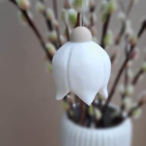 May include: A white, bell-shaped ornament with a light brown top, suspended in front of a blurred background of pussy willow branches. The ornament hangs from a thin string. A white vase is visible at the bottom.