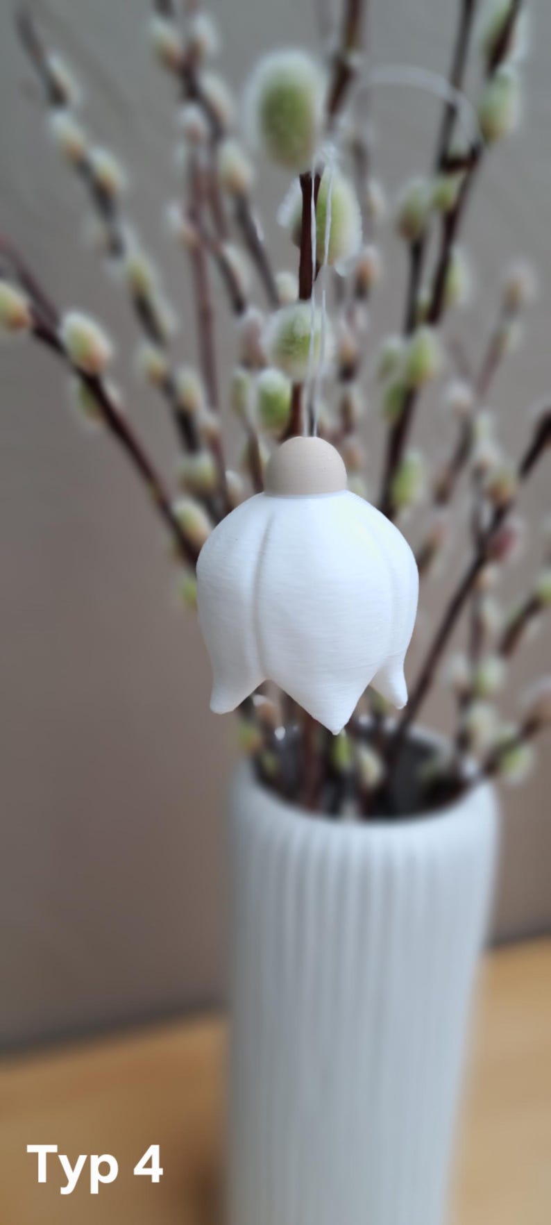 May include: A white, bell-shaped ornament with a light brown top, suspended in front of a blurred background of pussy willow branches. The ornament hangs from a thin string. A white vase is visible at the bottom.