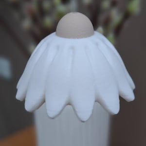 May include: A white, daisy-shaped decorative object with a tan center and a white vase. The object is labeled "Typ 1". The background is blurred, featuring a glimpse of greenery.