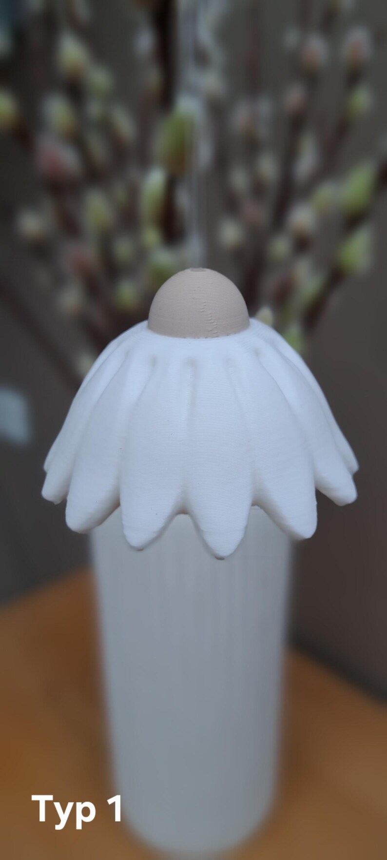 May include: A white, daisy-shaped decorative object with a tan center and a white vase. The object is labeled "Typ 1". The background is blurred, featuring a glimpse of greenery.