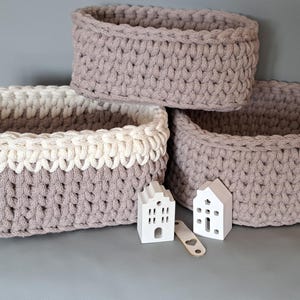 Könnte beinhalten: Drei handgehäkelte Körbe in verschiedenen Größen. Zwei sind einfarbig taupe, und einer hat einen cremefarbenen oberen Rand. Kleine weiße hausförmige Dekorationen und ein Holzanhänger befinden sich vor den Körben.