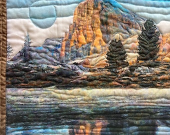 May include: A quilted wall hanging featuring a landscape scene with a mountain, trees, and a lake. The mountain is a vibrant orange and yellow, while the lake is a deep blue with a reflection of the mountain. The sky is a light blue with white clouds.