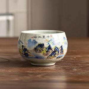 May include: A small, off-white ceramic tea cup with a landscape design. The cup features a mountain and water scene in blue, yellow, and green. The rim has Chinese characters. The cup sits on a wooden surface.