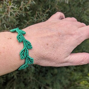 May include: A green macrame bracelet with a leaf pattern. The bracelet, made from knotted cord, is displayed on a wrist. The background shows blurred green foliage.