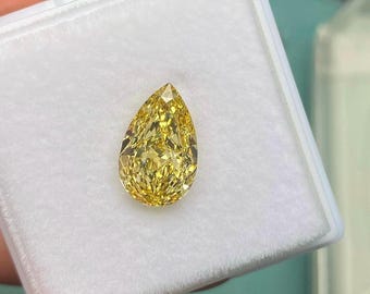 May include: A pear-shaped, fancy vivid yellow diamond gemstone. The diamond is set in a white box, and the facets reflect light, creating a brilliant sparkle. The gemstone is held by a hand.