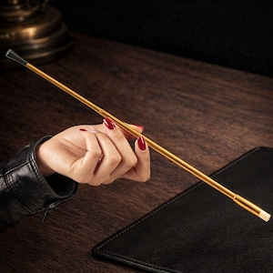 May include: A gold-colored cigarette holder with a black tip and a white end. The slender holder is held in a hand with red nail polish, resting on a dark wooden surface. A black leather-like item is in the background.