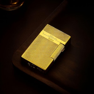 Puede incluir: Un encendedor dorado con una superficie texturizada con patrón de diamantes. El encendedor tiene forma rectangular con una tapa con bisagras y una pequeña sección rectangular con líneas verticales. La marca es visible.