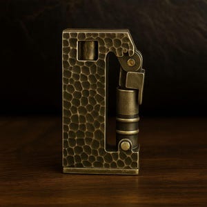 Op de afbeelding: Een bronzen aansteker in vintage-stijl met een getextureerde, gehamerde afwerking. De aansteker heeft een rechthoekige behuizing met een scharnierend deksel en een cilindrische brandstofkamer. Het ontwerp heeft een unieke, industri&euml;le esthetiek.