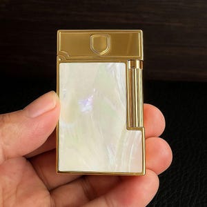 May include: A gold-coloured lighter with a rectangular shape and a shield emblem on the top. The lighter has a white, iridescent panel on the front. The design includes vertical gold stripes on the side.