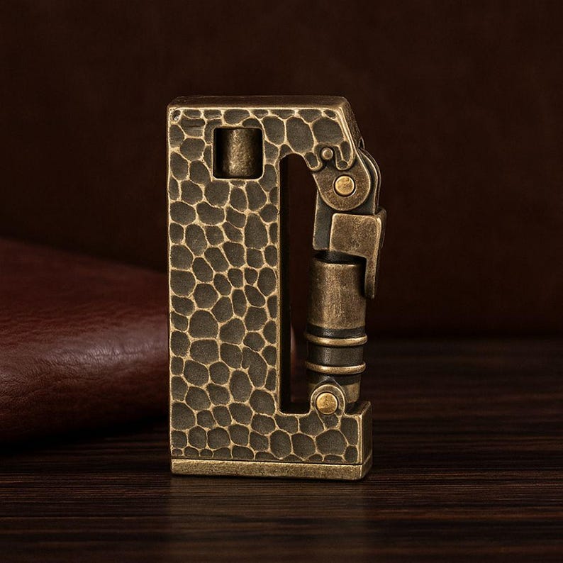 Op de afbeelding: Een vintage messing aansteker met een getextureerd, gehamerd oppervlak. De rechthoekige behuizing heeft een scharnierend deksel en een gedetailleerd mechanisme. De antieke messingkleur suggereert een klassieke of steampunk esthetiek.