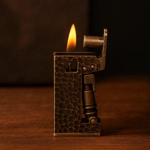 Op de afbeelding: Een vintage-stijl aansteker met een getextureerde, donkerbronzen afwerking. De aansteker is rechthoekig met een scharnierend deksel en een mechanische hendel. Een helder oranje vlam is zichtbaar, wat aangeeft dat de aansteker in gebruik is. Het ontwerp suggereert vakmanschap en een klassieke esthetiek.