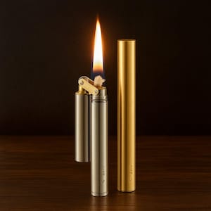 May include: A lit, silver and brass lighter with a long, orange flame. A closed, gold-colored case stands next to the lighter. The lighter is open, revealing a wick. The lighter and case are on a wooden surface.