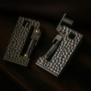 Op de afbeelding: Twee metalen aanstekers met een getextureerde, gehamerde afwerking. De aanstekers zijn donkergrijs met zwarte en gouden accenten. E&eacute;n aansteker is open en onthult de interne componenten. De andere aansteker is gesloten.