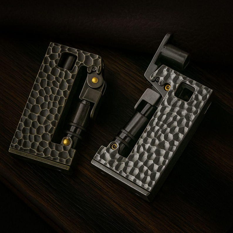 Op de afbeelding: Twee metalen aanstekers met een getextureerde, gehamerde afwerking. De aanstekers zijn donkergrijs met zwarte en gouden accenten. E&eacute;n aansteker is open en onthult de interne componenten. De andere aansteker is gesloten.