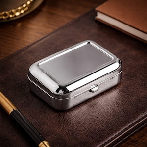 May include: A rectangular, silver-colored pill box with a shiny, reflective surface. The box has a hinged lid and a small clasp. It rests on a brown leather surface, with a gold and black pen in the foreground.