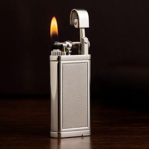 May include: A silver lighter with a lit flame. The rectangular body has a textured, diagonal-lined panel. The top has a hinged cover and ignition mechanism. The flame is orange and yellow. The lighter is on a dark surface.