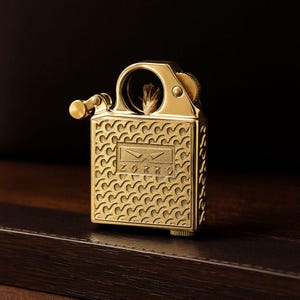 Puede incluir: Un encendedor ZORRO de color dorado con un intrincado patrón de ondas. El encendedor tiene un cuerpo rectangular con una tapa con bisagras y una pequeña rueda redonda de ajuste de combustible. El logotipo de ZORRO se muestra en la parte delantera.