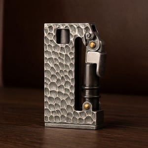 Op de afbeelding: Een zilverkleurige, getextureerde metalen aansteker met een uniek ontwerp. De aansteker heeft een gehamerd oppervlak op de hoofdbehuizing en een scharnierende bovenkant met een cilindrische brandstofkamer. Kleine goudkleurige accenten voegen details toe.
