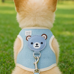 Puede incluir: Un arnés azul claro para perros con un diseño de cara de oso. El arnés tiene un ribete color crema y un cierre metálico. Las palabras "HAND MADE" están impresas en el arnés. El perro es de color marrón claro y está sentado sobre hierba verde.