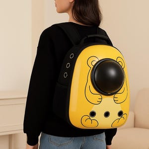 May include: Yellow pet carrier backpack with a black bubble window and bear cartoon design. The backpack has black straps and a handle. The carrier has three small air holes and is worn by a person wearing a black sweater and blue jeans.