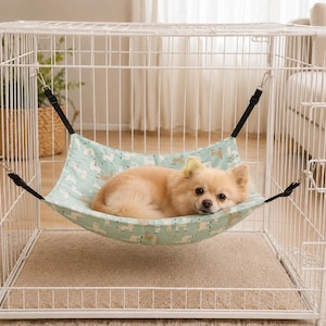 May include: A light green fabric hammock featuring a repeating white llama pattern, suspended by four black adjustable straps with metal clips inside a white wire pet crate, with a small tan dog resting comfortably on the fabric surface.