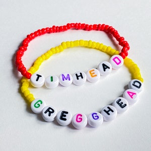 Puede incluir: Dos pulseras de cuentas con los nombres "TIM HEAD" y "GREG HEAD" deletreados en letras de colores. Las pulseras están hechas con cuentas blancas y hilo rojo, amarillo y negro.