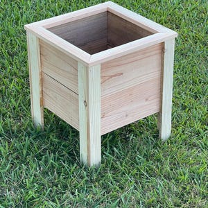 Könnte beinhalten: Ein quadratischer Pflanzkasten aus Holz mit natürlicher Holzoberfläche. Der Pflanzkasten hat ein schlichtes Design mit vier Beinen und einem erhöhten Rand. Er steht auf einer grünen Wiese.