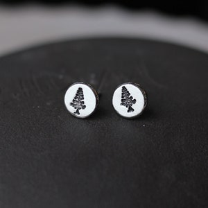 May include: A pair of silver stud earrings with a tree design. The earrings are round with a white background and a black tree silhouette. They are displayed on a dark, textured surface.