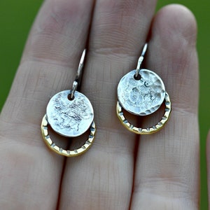 May include: A pair of silver and gold earrings with a hammered texture. The earrings are made of two circles, one larger than the other, with the smaller circle sitting on top of the larger circle. The earrings are hanging from a silver hook.