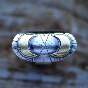 May include: A silver ring featuring two gold crescent moons on the top. The ring has a textured, hammered finish.