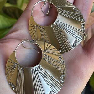 May include: A pair of silver hoop earrings with a sunburst design and leaf accents.