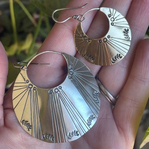 May include: A pair of silver hoop earrings with a sunburst design and leaf accents. The earrings are handmade and have a unique, bohemian style.
