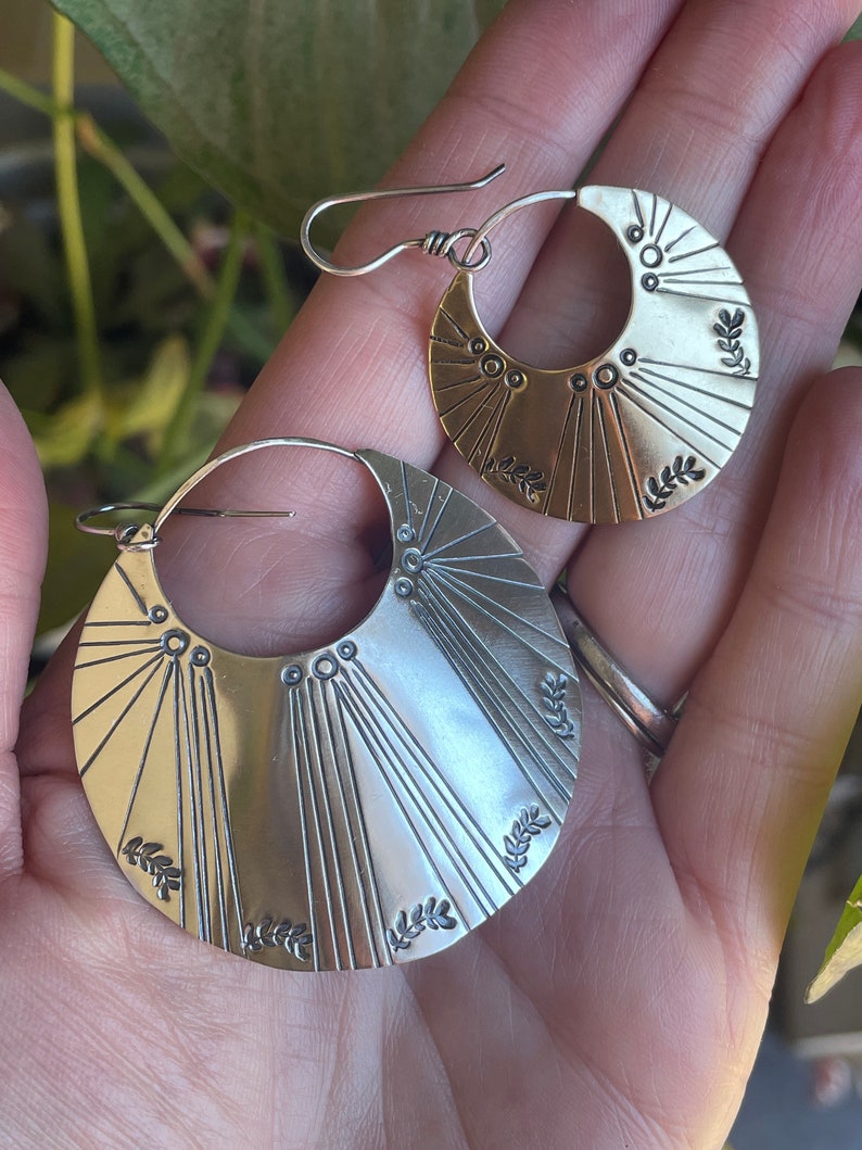 May include: A pair of silver hoop earrings with a sunburst design and leaf accents. The earrings are handmade and have a unique, bohemian style.
