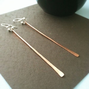 May include: A pair of long, thin copper earrings with silver hooks and beads. The earrings are hanging on a brown surface.