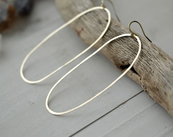 Può includere: Un paio di orecchini a cerchio ovali color oro. Gli orecchini sono allungati e hanno un design semplice e minimalista. Sono esposti su un pezzo di legno alla deriva su uno sfondo grigio chiaro.