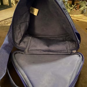 May include: Open view of a blue triangular-shaped bag with a zipper closure. The bag has a shoulder strap and a soft, padded interior. A small tag is visible inside the bag.