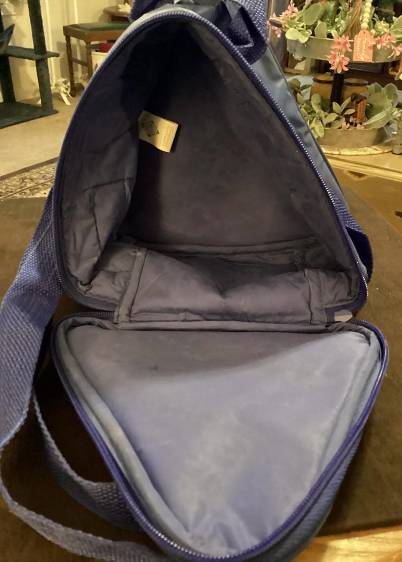 May include: Open view of a blue triangular-shaped bag with a zipper closure. The bag has a shoulder strap and a soft, padded interior. A small tag is visible inside the bag.
