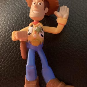 Pu&ograve; includere: Una piccola statuetta in plastica di un personaggio cowboy con cappello marrone, camicia gialla a maniche lunghe con gilet maculato, stella da sceriffo dorata, pantaloni blu e stivali marroni. La figura ha un braccio alzato e una fondina.