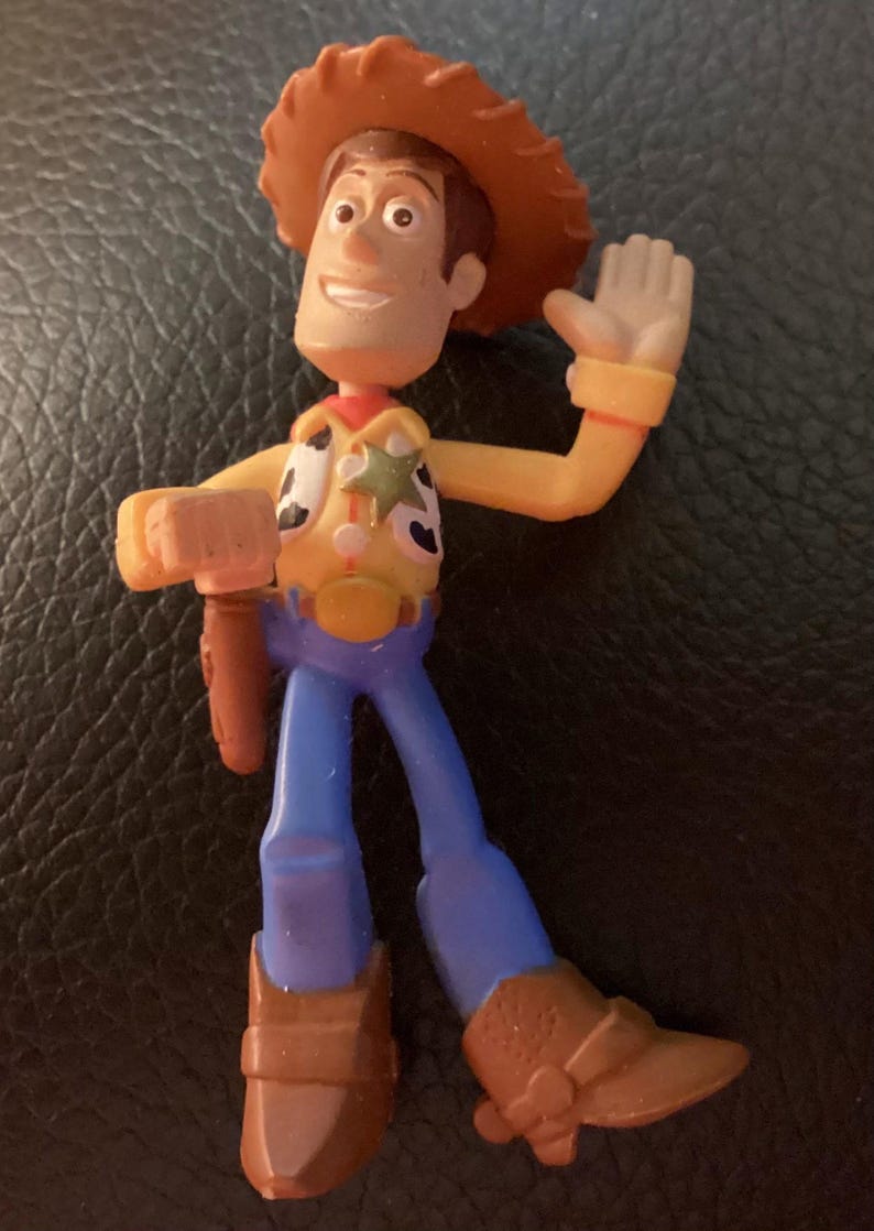 Pu&ograve; includere: Una piccola statuetta in plastica di un personaggio cowboy con cappello marrone, camicia gialla a maniche lunghe con gilet maculato, stella da sceriffo dorata, pantaloni blu e stivali marroni. La figura ha un braccio alzato e una fondina.