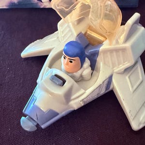 May include: A white and light blue toy spaceship with a clear cockpit. A small figure with a blue helmet and orange face is seated inside. The toy has wings and a blue nose. The background is a dark purple surface.
