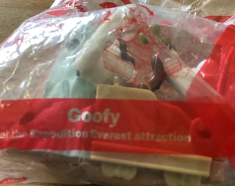 May include: A clear plastic bag containing a Goofy toy from the Expedition Everest attraction. The toy appears to be a small figurine with multiple colors. The bag has a red banner with the word "Goofy" printed on it.