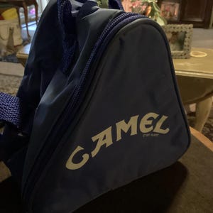 May include: A navy blue triangular bag with a zipper closure and a handle. The bag features the word "CAMEL" in a stylized font. The bag is made of a durable material and has a simple design.