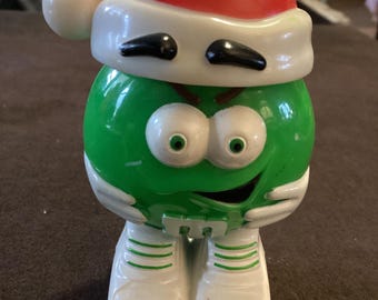 May include: A green candy character wearing a red Santa hat with a white brim and a white pom-pom. The character has white shoes with green stripes and black eyes. The figure has a smiling face and is a Christmas-themed collectible.
