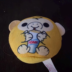 May include: A round, yellow plush toy featuring a cartoon monkey with light-colored ears. The monkey is holding a blue and white cup with the red text SONIC printed on it. A white fabric tag is attached to the bottom right side of the plush toy.