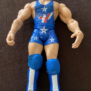 May include: A muscular action figure of a wrestler in a blue singlet and shorts with white stars. The figure has blue knee pads and boots with white laces. The singlet features a red and white eagle design.