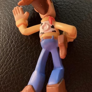 Pu&ograve; includere: Una piccola statuetta in plastica di un personaggio cowboy che indossa un cappello marrone, una camicia gialla con macchie nere, un fazzoletto rosso, pantaloni blu e stivali marroni. La figura ha una fondina sul fianco sinistro e un anello sul retro.